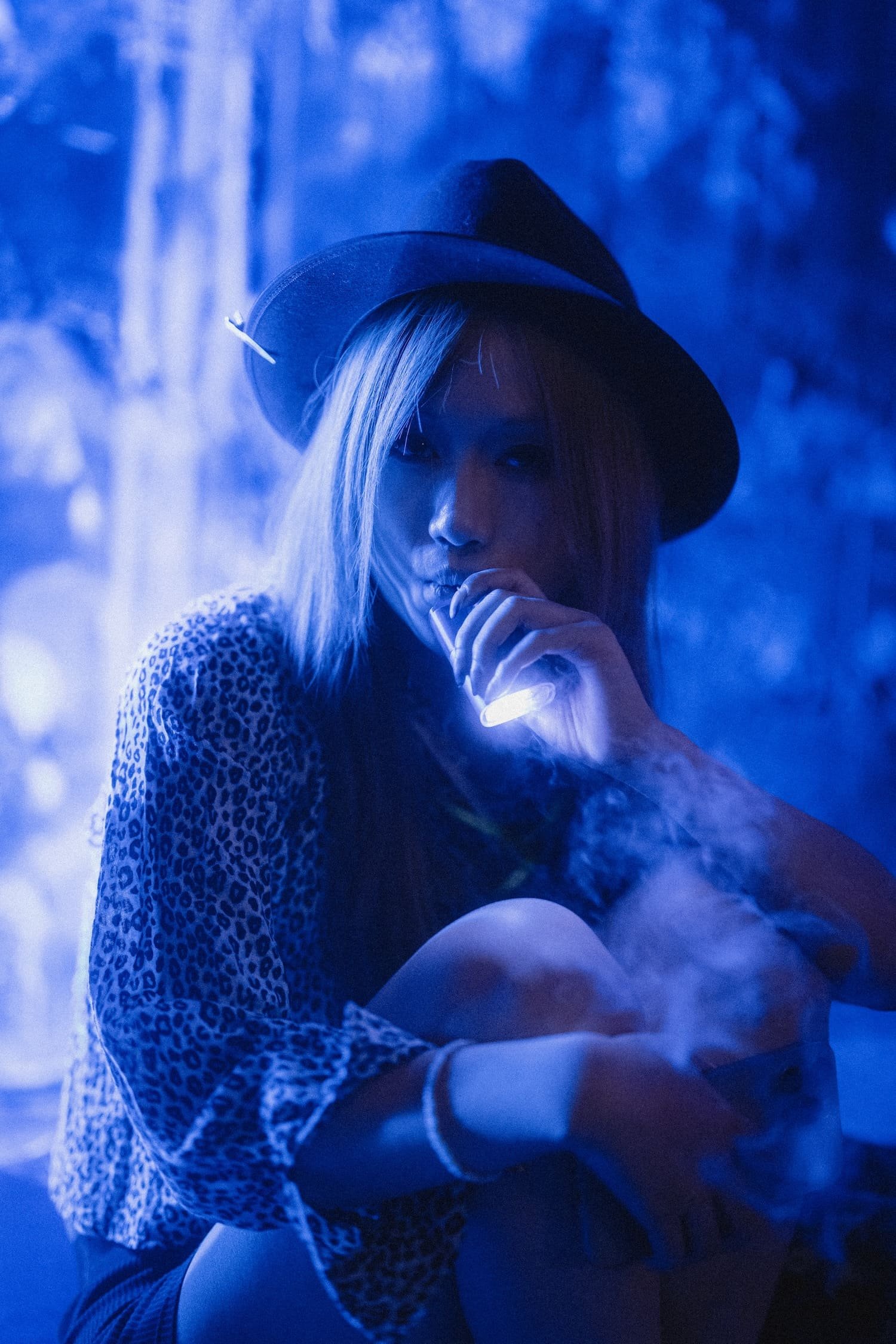 Bondage Ball 
---
A woman with long hair wearing a leopard print shirt and a wide-brimmed hat is holding a glowing object near her face, against a dark blue background with blurred trees.