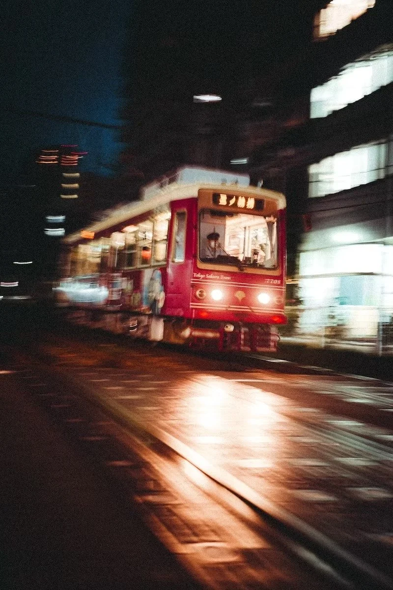 A red tram moving along tracks at night with blurred city lights in the background in Tokyo at night by Deniz Demir.