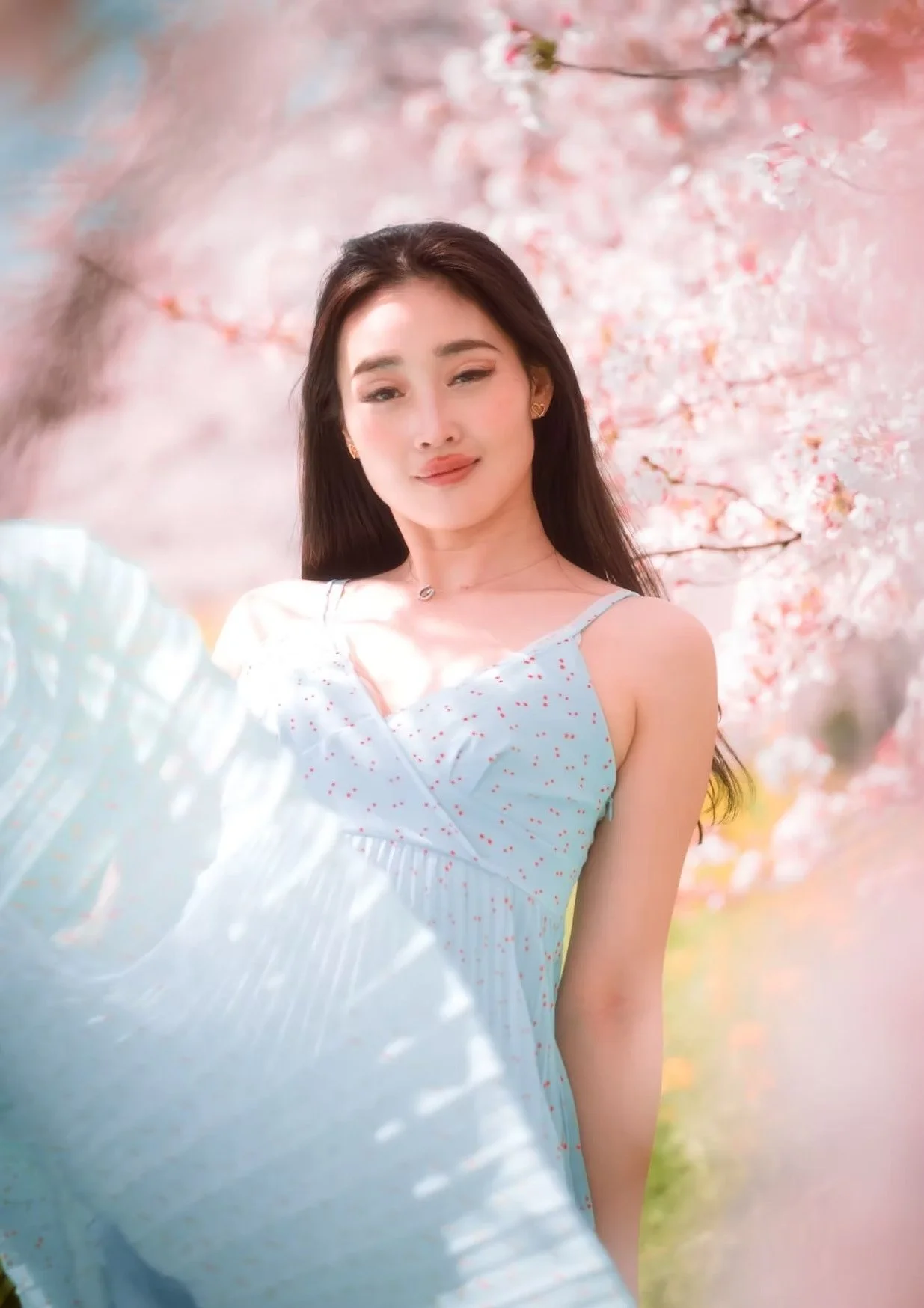 A woman in a light blue dress smiles as she twirls in a cherry blossom garden, with flowers surrounding her. The soft breeze and gentle sunlight add a dreamy, elegant touch to the scene.