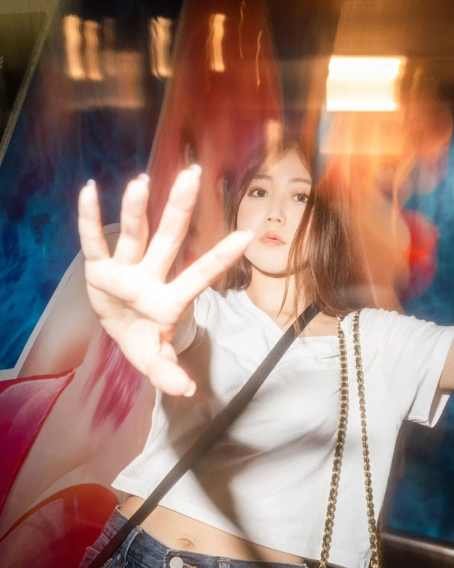 A woman with long brown hair, wearing a white top with a chain strap purse, is reaching out her hand with fingers spread, as she looks at her reflection behind a glass surface in a colorful environment.