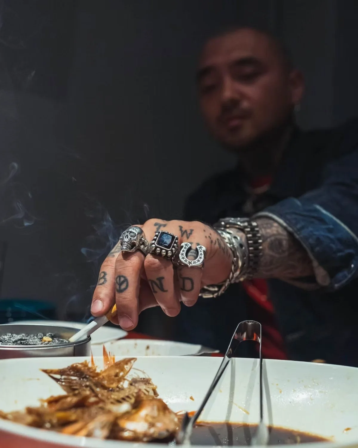 A Japanese man with tattoos on his hand and wrist, wearing rings and jewelry, is reaching into a bowl of food with a cigarette in his hand. He has tattoos on his fingers spelling 'T W D' and tattoos on his arm, and is dressed in a denim jacket.