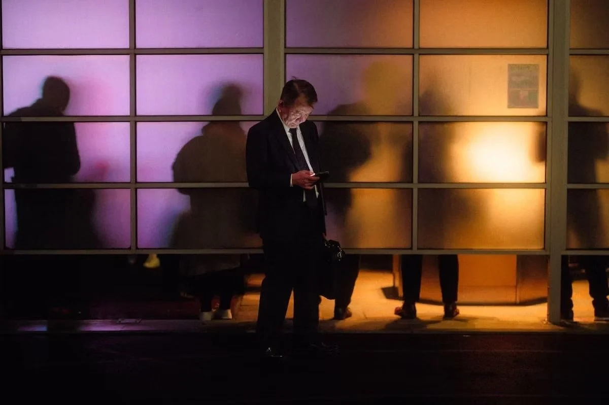 A Japanese salaryman in a suit standing outside a building at night, looking at his phone, with shadows of people behind a glass wall illuminated by warm and purple lighting. Shot by Tokyo street photographer Deniz Demir on the streets of Shimbashi.