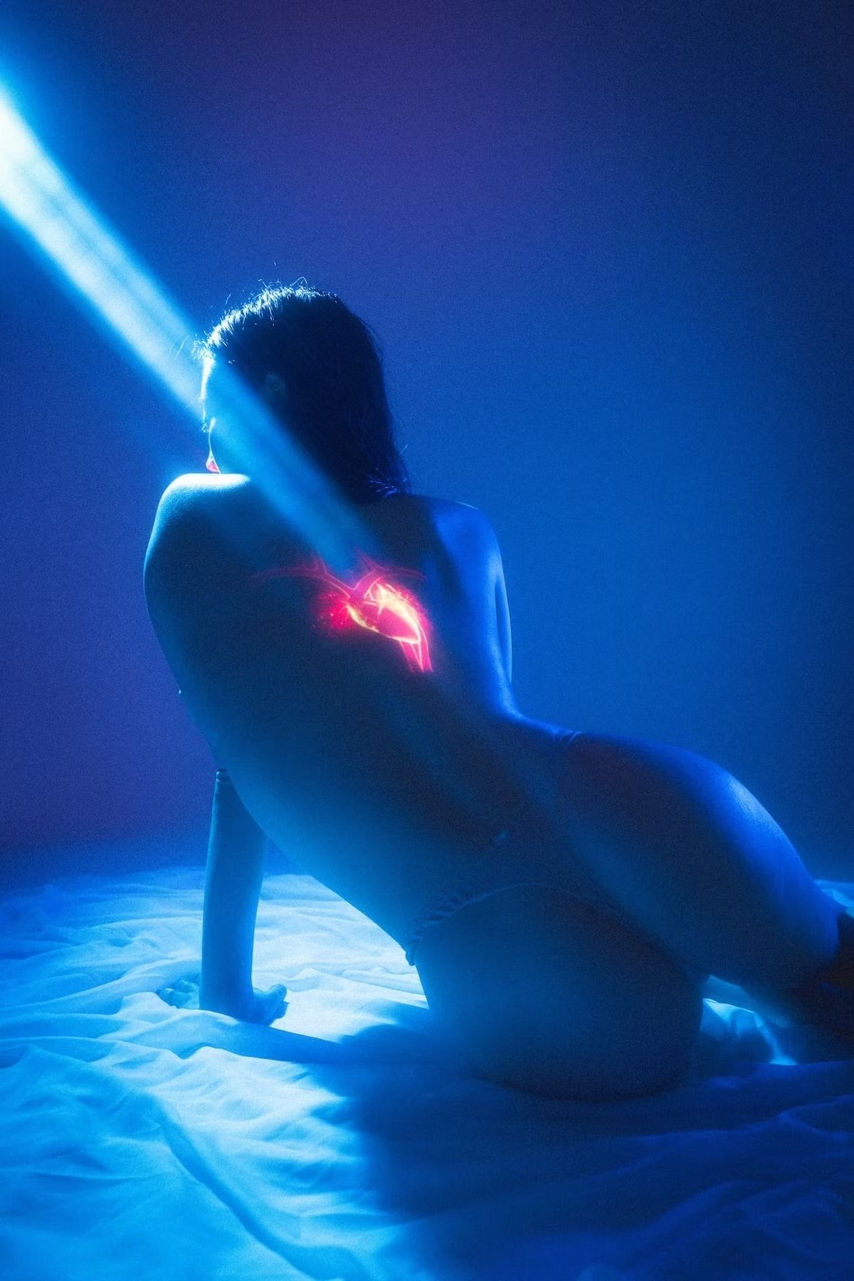 A woman with short hair kneeling on a white surface in front of a dark blue background illuminated by a blue light. She is topless and has a glowing red and pink heart shape projected onto her chest, with the shape radiating light.