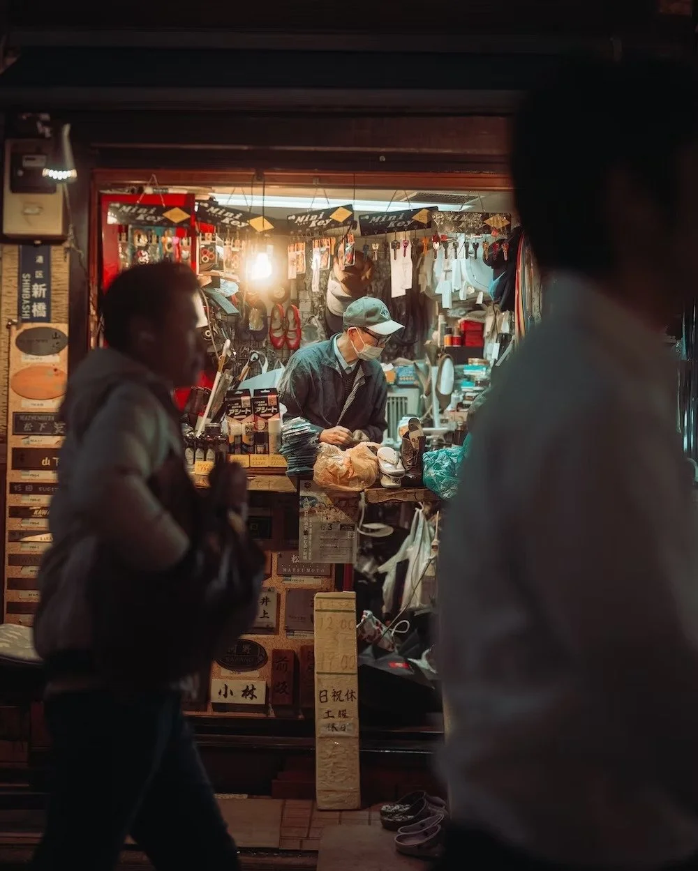A street scene showing a small shop with a man behind the counter wearing a face mask and cap, selling various goods. In the foreground, there are two pedestrians walking past the shop, slightly out of focus.