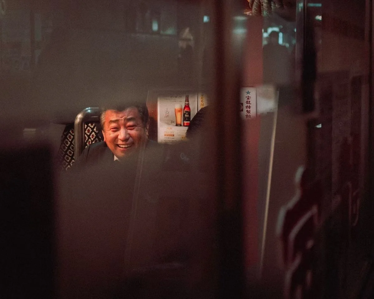 A Japanese salaryman with a big smile sitting at a table in a dimly lit restaurant or bar, seen through a partially obscured or reflective surface.