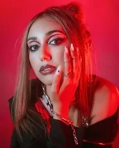 A young woman with long hair and makeup, wearing a black top, touching her face with her hand, against a red background.