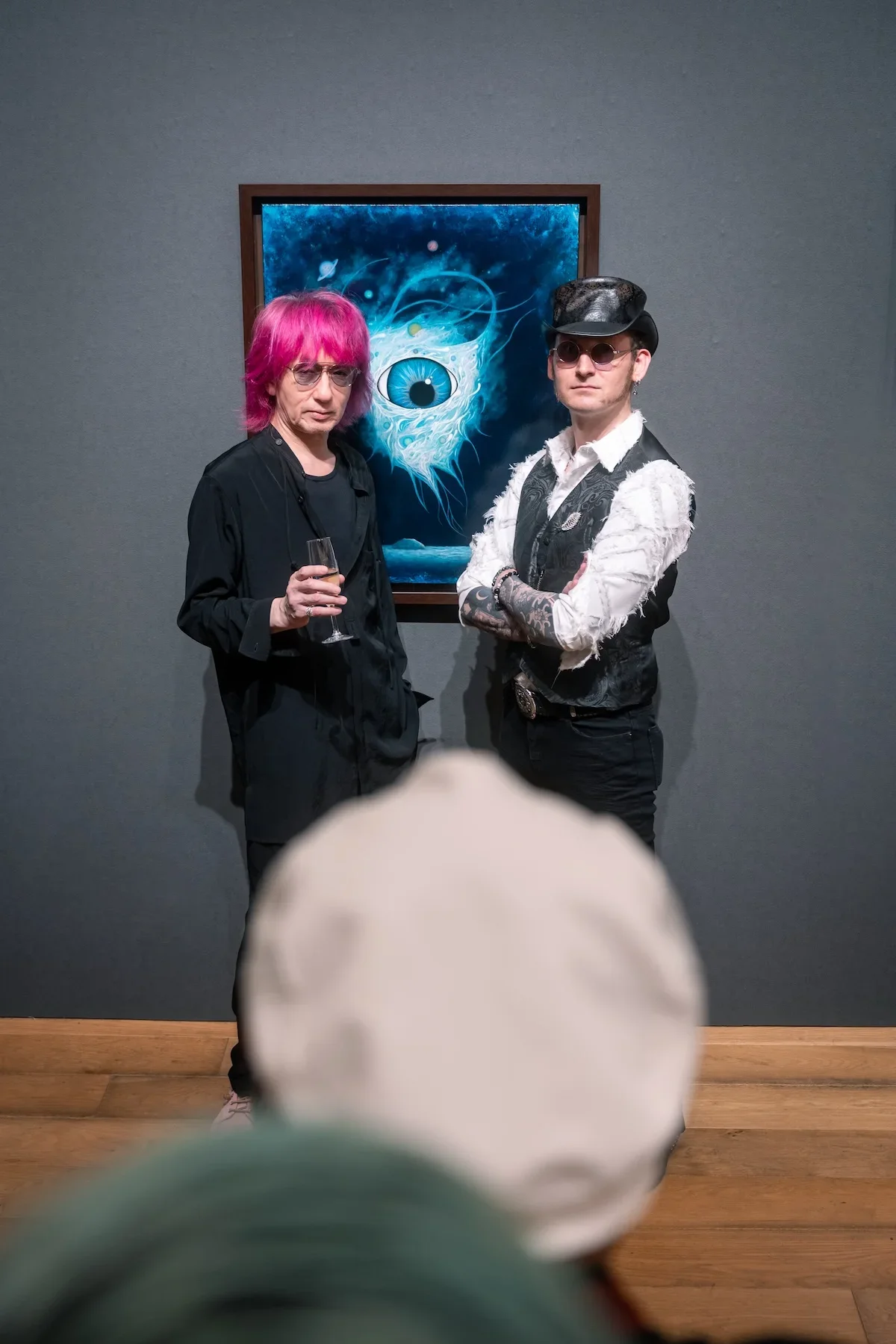 August Vilella
---
Two guests pose in front of a blue-eyed surrealist creature painting at August Vilella's art exhibition, surrounded by visitors enjoying the contemporary artwork and gallery atmosphere.