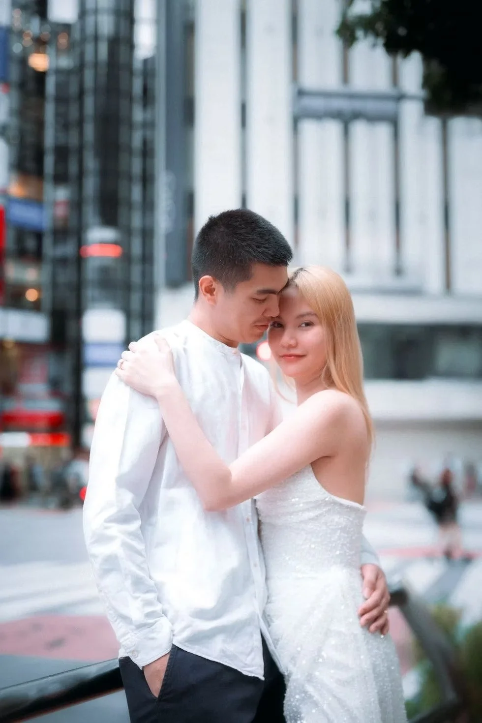 A couple dressed in white outfits embraces each other in an urban setting in Tokyo, their backs to tall buildings and bustling streets.