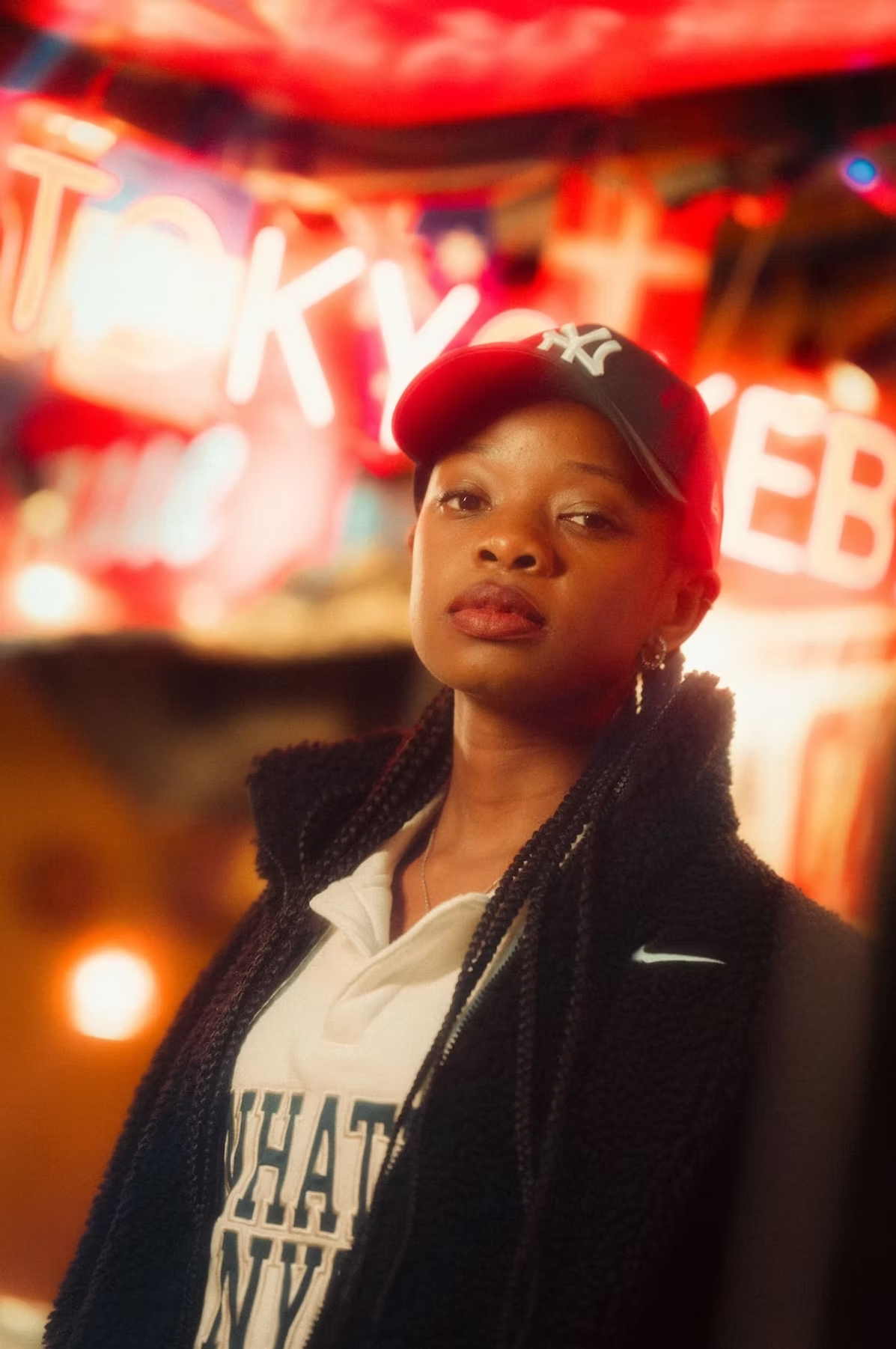 A woman wearing a white shirt, black jacket, and a New York Yankees cap stands in front of blurred neon lights in Tokyo, with a focused expression.