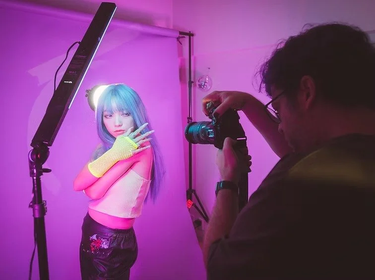 A photographer Daniz damir taking a picture of a model with blue hair posing under pink and purple lighting in a studio.