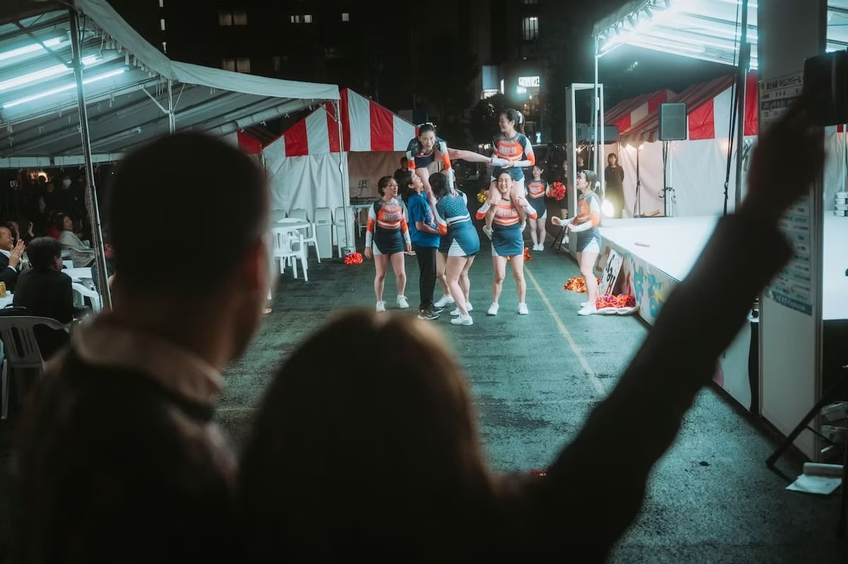 Cheerleaders performing a routine at a nighttime outdoor event with an audience seated under tents.