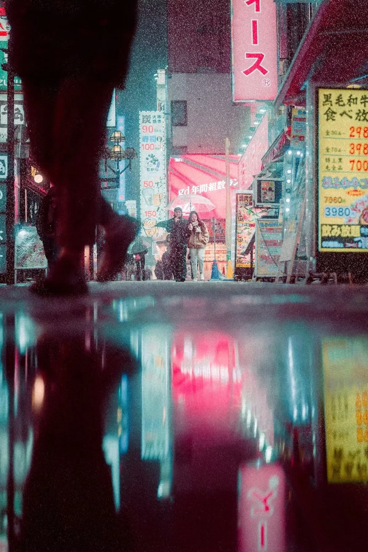 A rainy night scene on a city street in Japan, viewed from low angle near the ground. People are walking with umbrellas, illuminated signs and advertisements are visible, and the pavement reflects the colorful lights.