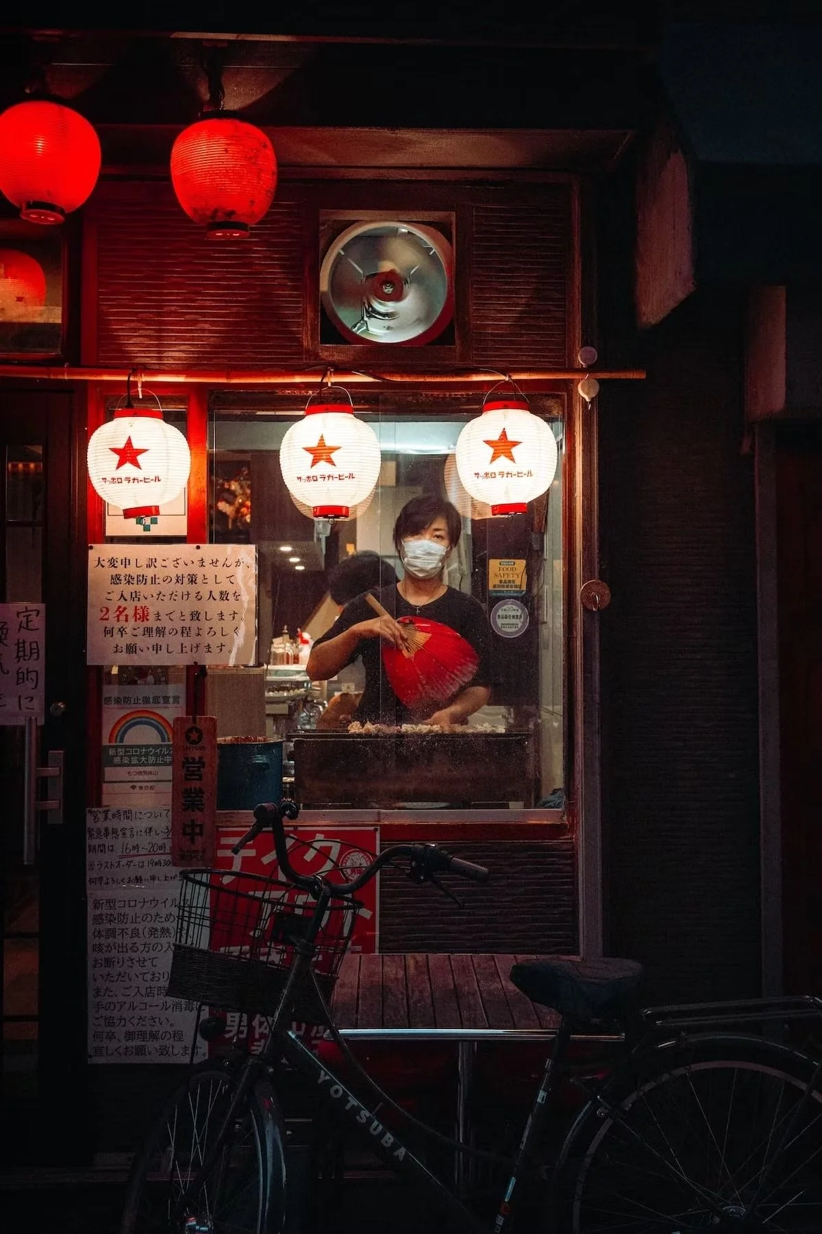 A Japanese street food stall with red lanterns and a woman wearing a face mask preparing food inside, viewed through a glass window. Bicycles are parked in front of the stall.