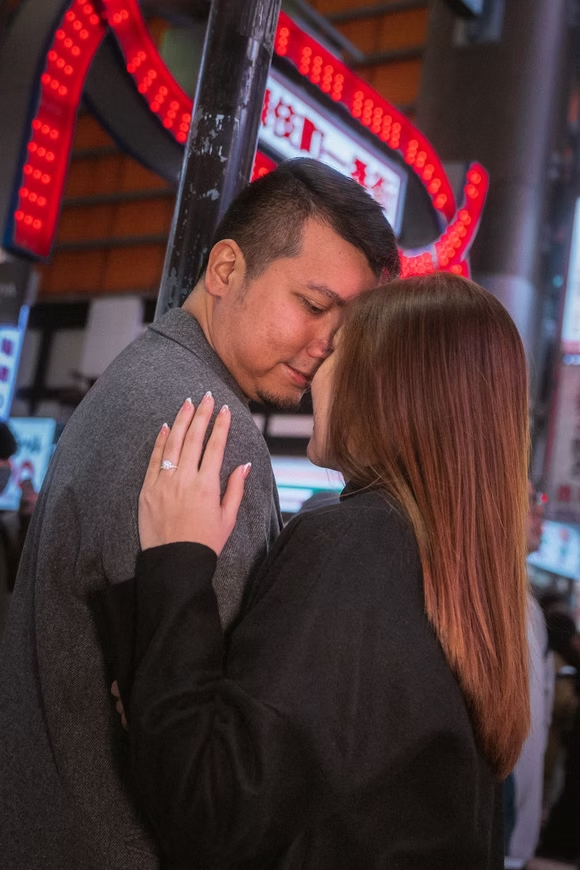A man and woman sharing an intimate moment, close to each other with their foreheads touching, in a lively urban setting with bright lights and signs in the background.