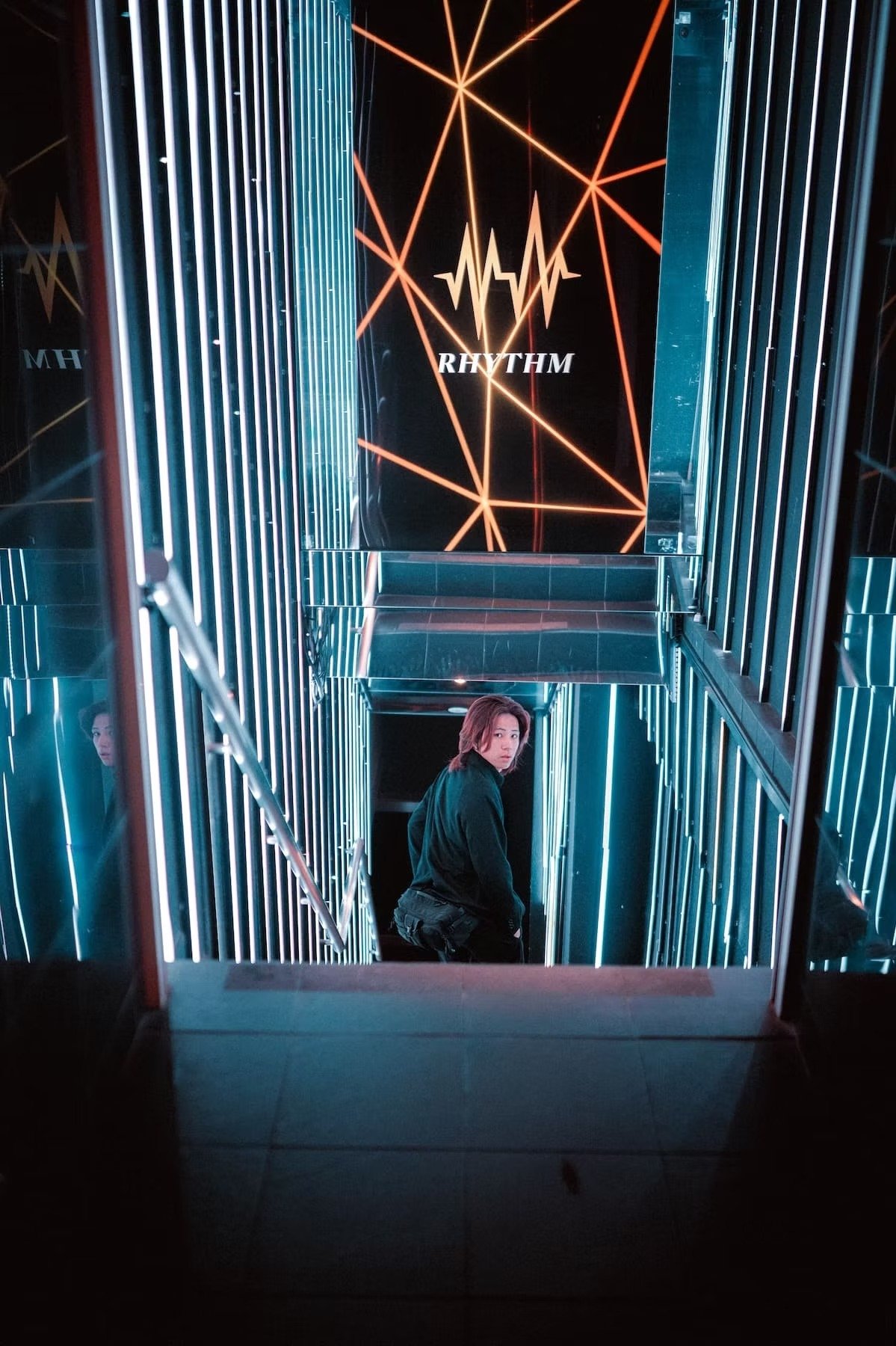 A Japanese host in Kabukicho with shoulder-length hair and a black jacket standing on a neon-lit staircase at night, with reflective mirrors on the sides and a black wall with orange neon lines and the word 'RHYTHM' in the background.