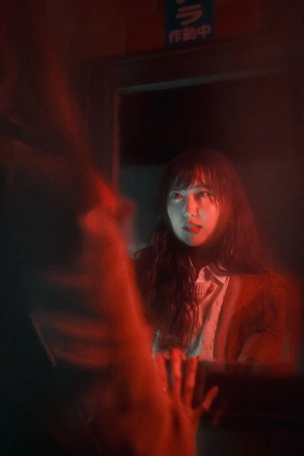A woman with long dark hair is illuminated by red lighting and looks in a specific direction, possibly at another person or object, in a dimly lit setting.