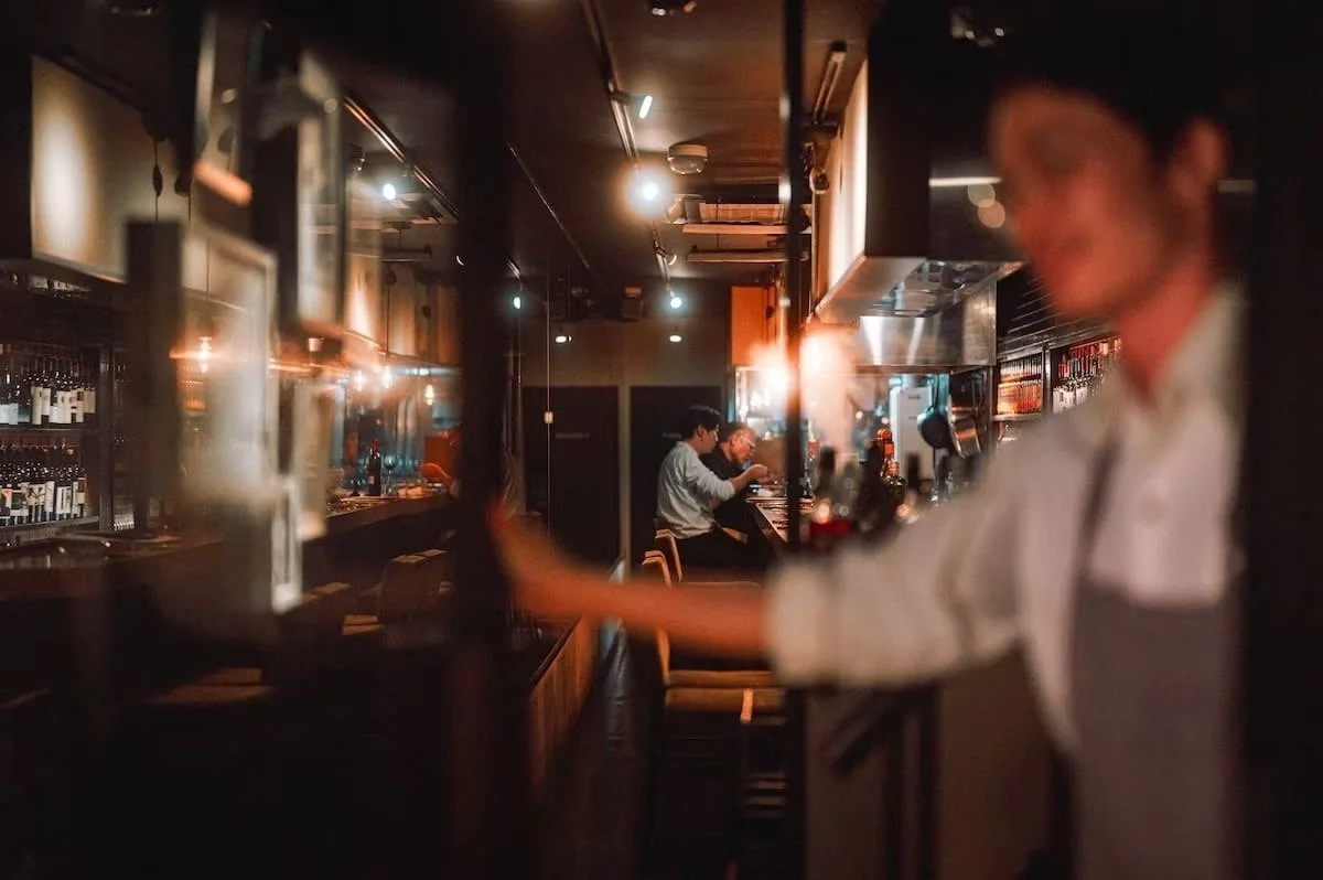 A blurry bar scene with people sitting on stools at the counter, including a bartender in foreground on the right, and customers in the background, illuminated by warm lighting.