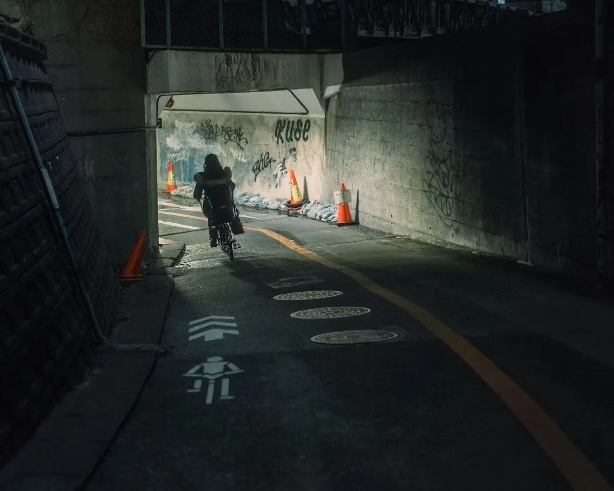 A person riding a bicycle through an underpass or tunnel with street art and graffiti on the walls. There are orange traffic cones and sandbags along the sides, and lane markings for bicycles and pedestrians.