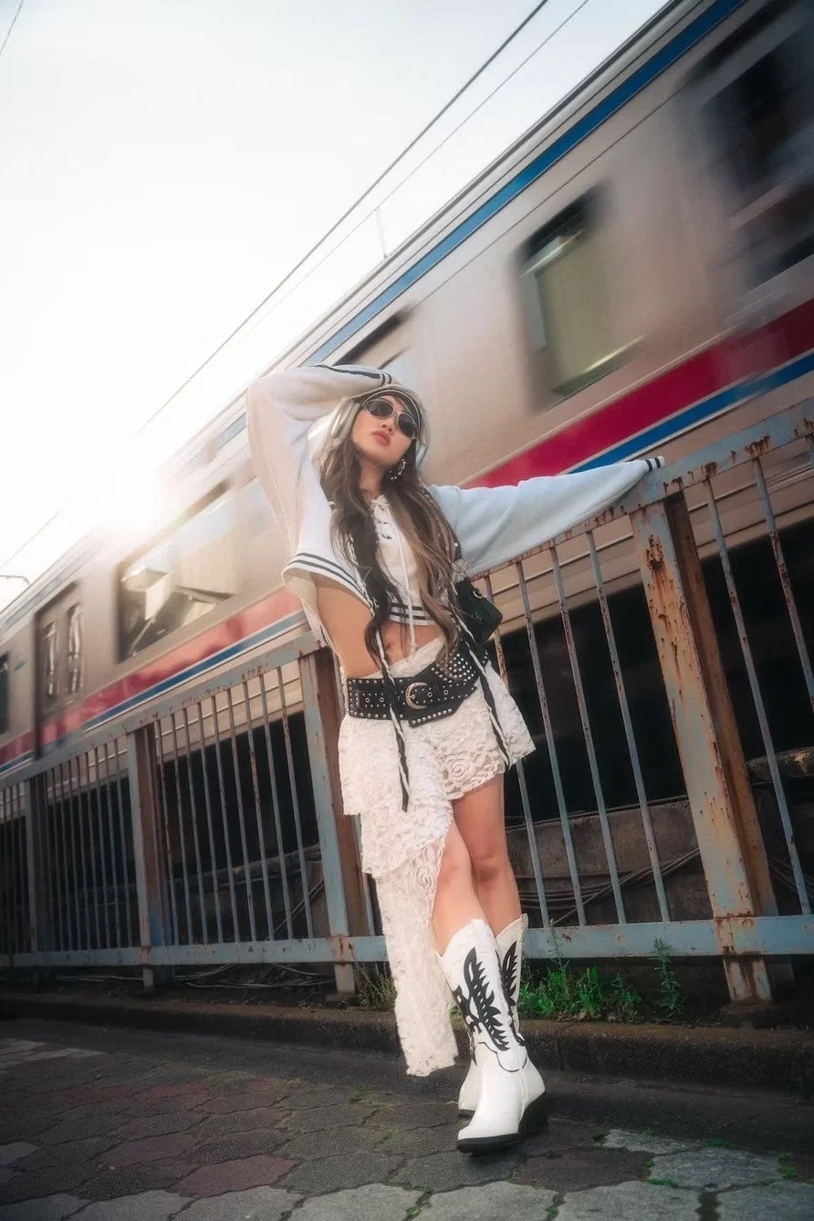 A woman in a white outfit with lace, black belt, and cowboy boots posing beside a railing with a moving train in the background in Tokyo Japan.