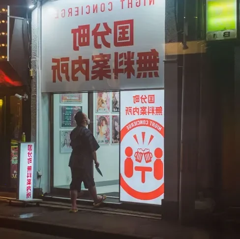 A person walks towards a brightly lit storefront with red and white signage at night, featuring Japanese text and images of people inside.