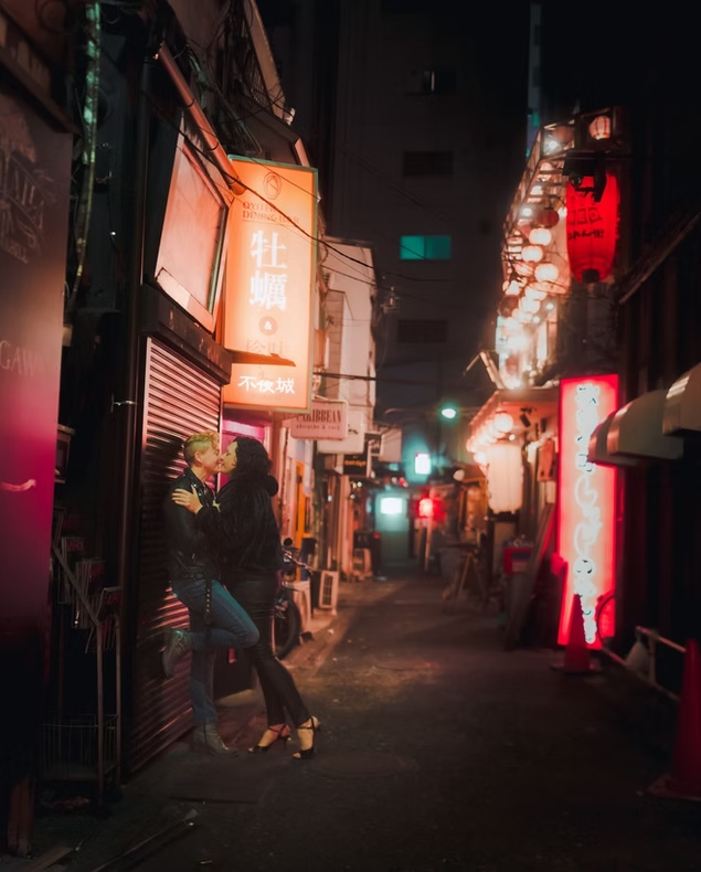 Two friends walk down a brightly lit alley in Tokyo at night, surrounded by colorful neon signs and glowing lights. One friend wears a leather jacket and the other smiles as they share a moment together.