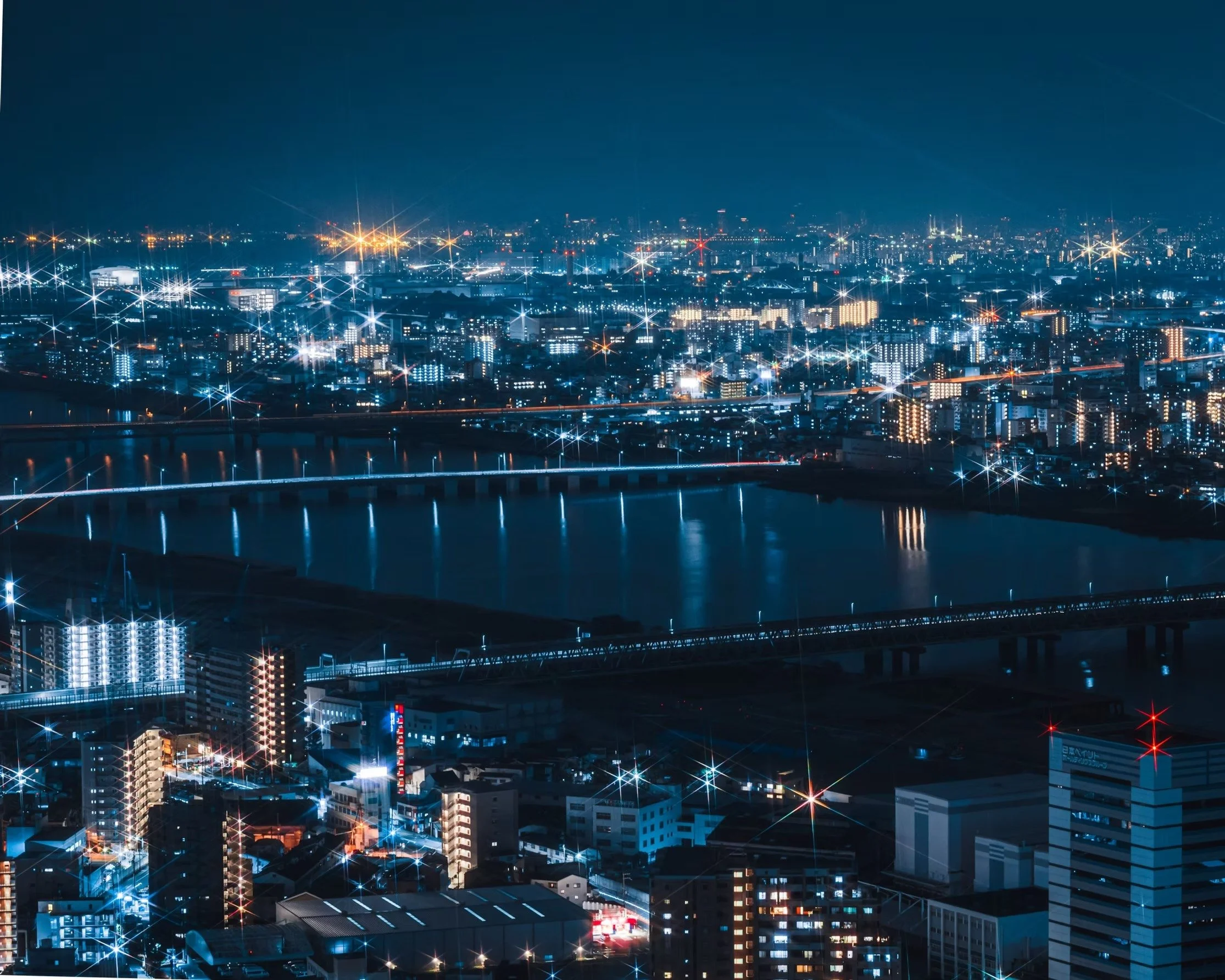 Nighttime cityscape with illuminated buildings and streets, reflecting on the water below, and a clear dark sky.