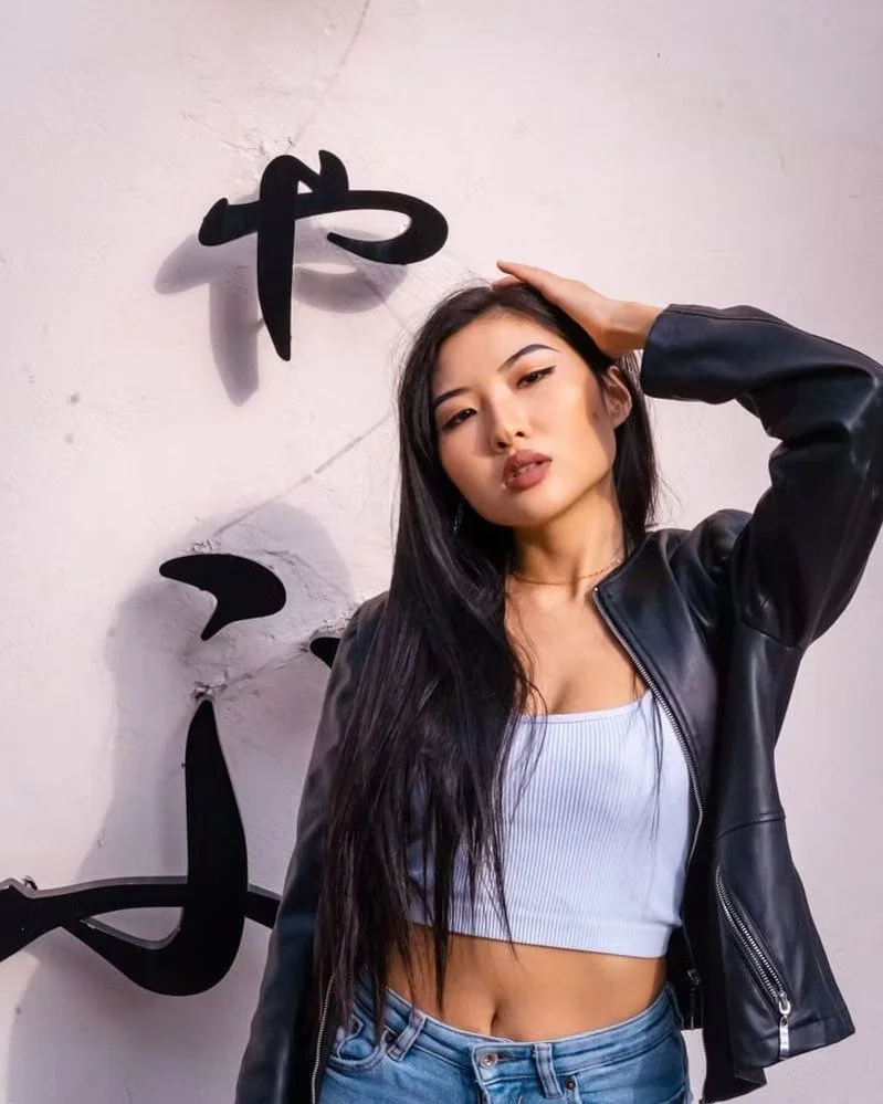 A young woman with long black hair, wearing a black leather jacket and a white crop top, posing with one hand on her head in front of a wall with Japanese characters.