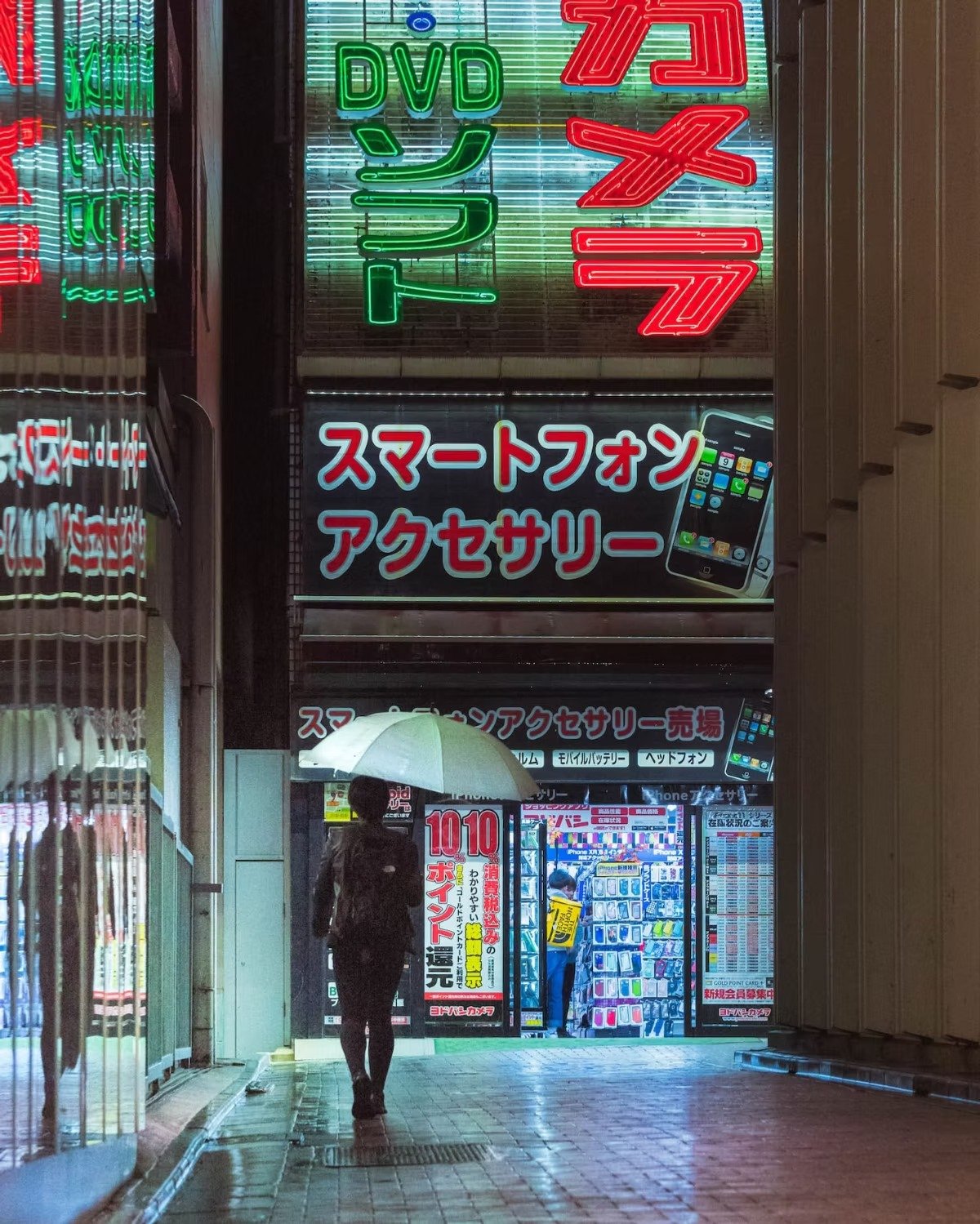 A Japanese person walking under an umbrella past a store with illuminated signs in Japanese advertising smartphones, accessories, and electronics, during a rainy night in a Tokyo alley.