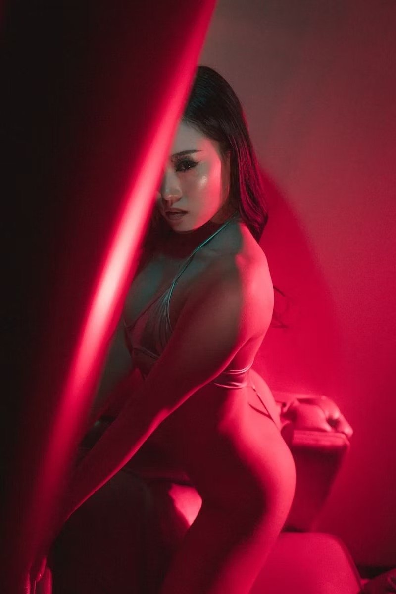 A woman with dark hair in a dimly lit room illuminated by red light, kneeling on a couch, wearing a revealing outfit, with a serious expression on her face.