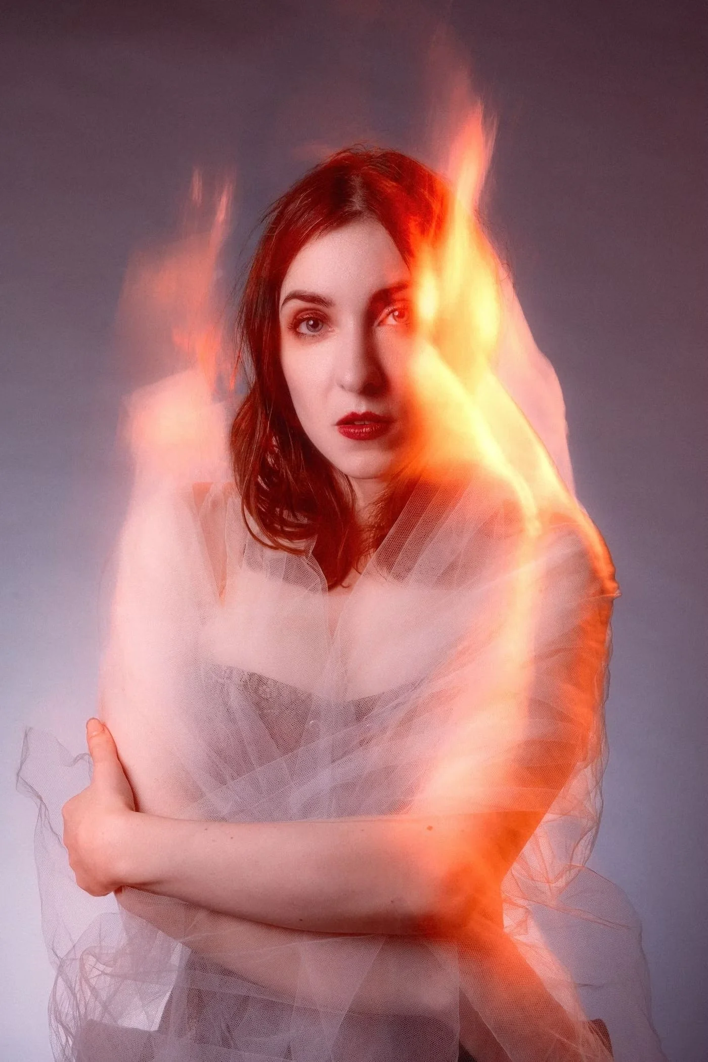 A woman with shoulder-length brown hair and red lipstick, partially obscured by glowing orange-red flames and wisps of sheer fabric, against a dark background.