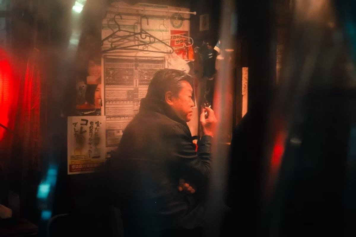 A man with dark hair wearing a black jacket, sitting in a dimly lit, Japanese, bar or restaurant, smoking a cigarette with posters and hangers on the wall behind him.