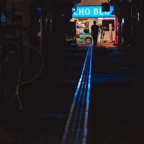 A dimly lit alley in Tokyo with a tactile paving strip glowing blue, leading to a brightly lit storefront where a silhouetted figure stands.