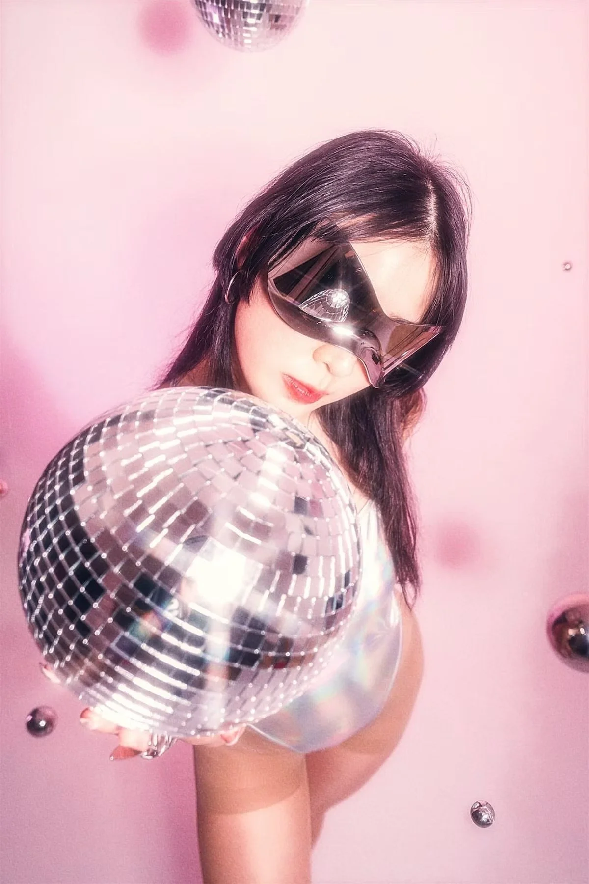 A woman with dark hair wearing large reflective sunglasses and holding a disco ball, standing against a pink background with smaller metallic balls.