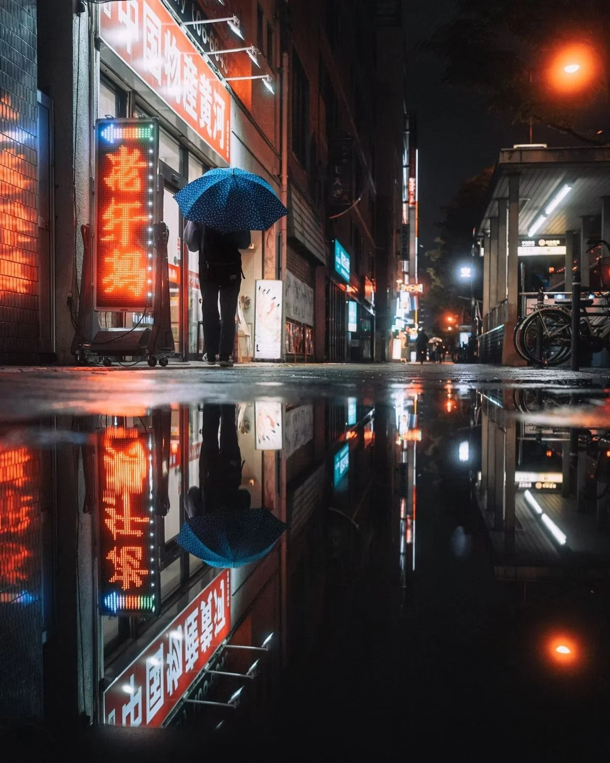 A person holding a polka-dotted umbrella walking on a wet city street at night, with neon signs reflected on the pavement, and bicycles parked on the sidewalk in Osaka, Japan.