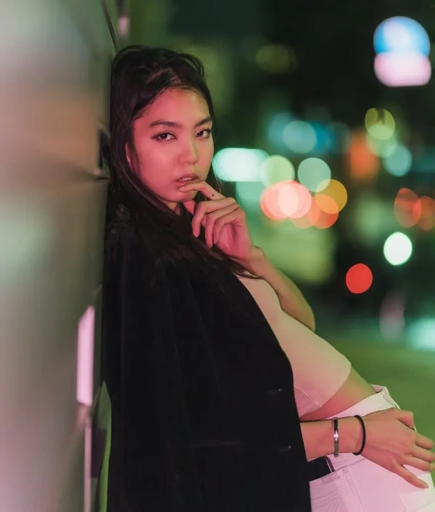 A young woman with dark hair leaning against a wall at night, with colorful city lights blurred in the background, gazing at the camera with her finger near her lips.