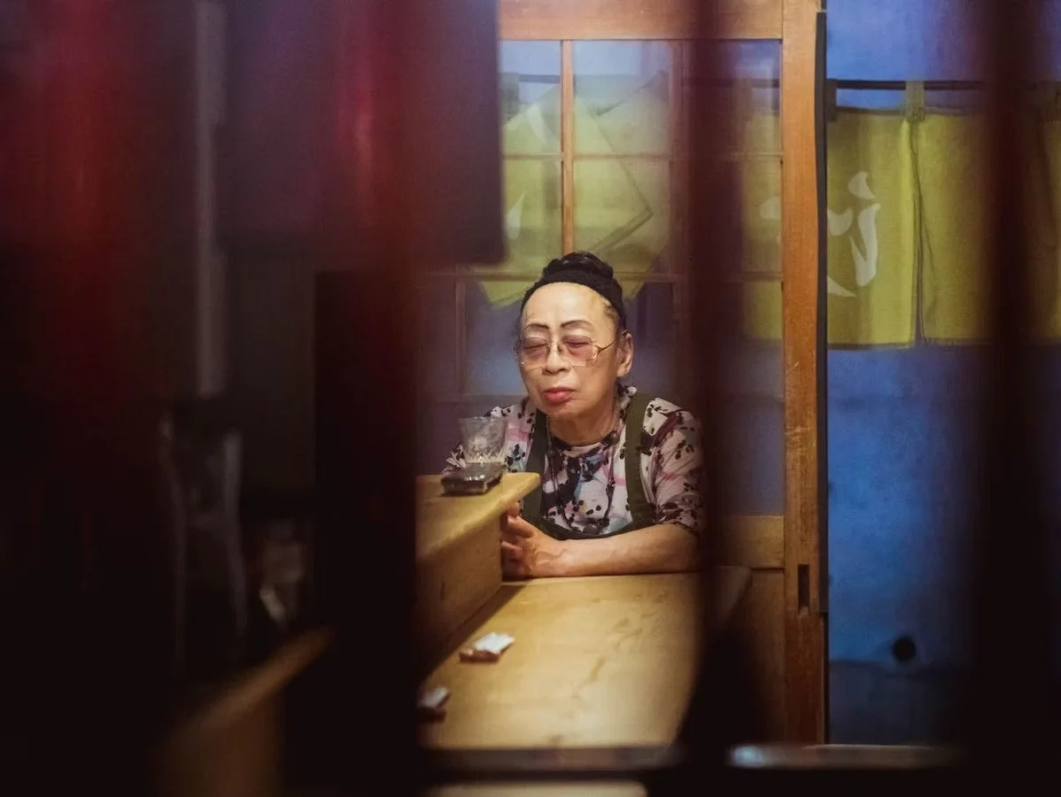 A Japanese woman sitting alone at a wooden bar, seen through a partially open door or partition, with her eyes closed and an expression of contemplation, wearing glasses, a patterned shirt, and a headband.