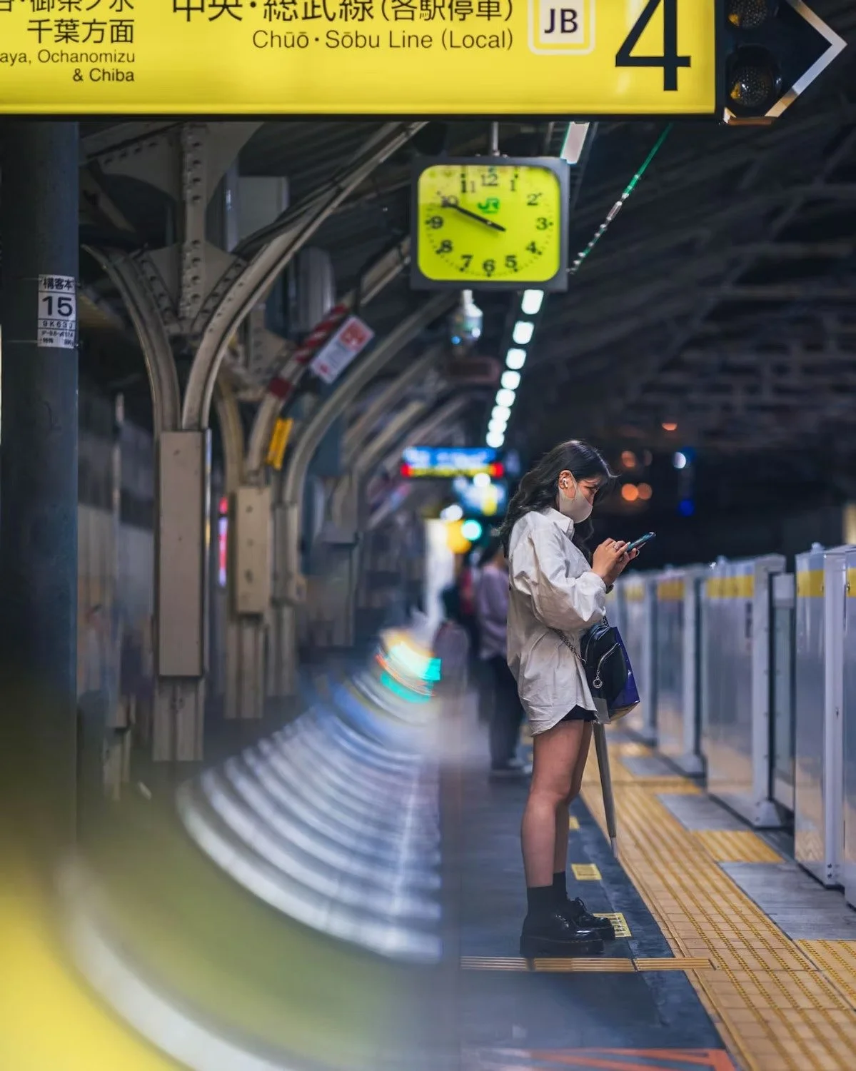 A woman standing on a train platform at night, looking at her phone while wearing a face mask. There are other people in the background, and a yellow clock showing the time at approximately 9:47. The platform is illuminated with overhead lights and s