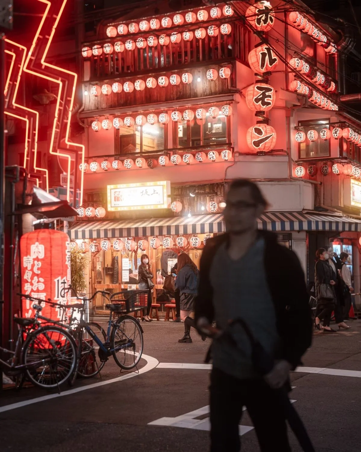 A bustling street scene at night in Japan with a multi-story building decorated with numerous red paper lanterns featuring Japanese characters. People are walking and dining outside, with bicycles parked nearby and neon signs illuminating the area. O