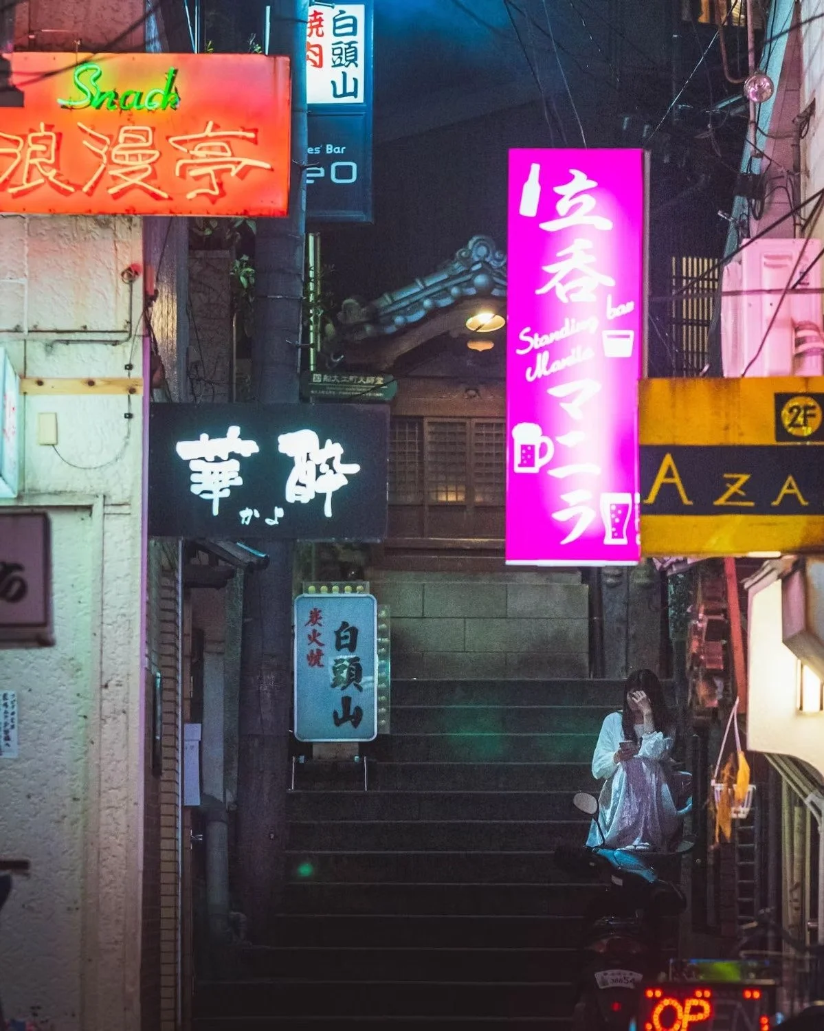 Street photography night scene in Nagasaki of a narrow street with multiple illuminated colorful signs in Japanese, including neon signs for bars and restaurants, and a woman sitting on a scooter while using her phone.