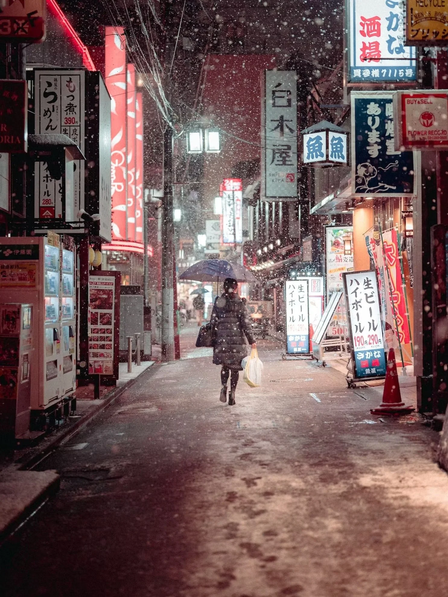 A person walking on a snowy street at night, holding an umbrella and carrying a plastic bag, surrounded by illuminated signs and storefronts with Japanese text.