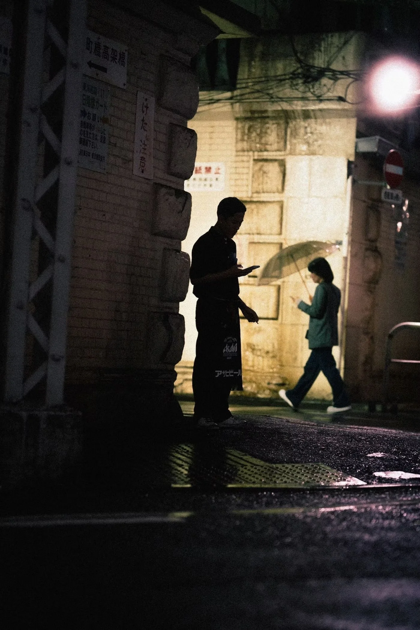 Nighttime street scene with two silhouetted people, a man and a woman, standing near a brick wall. The man is engrossed in his phone, while the woman holds an umbrella and walks past. The scene is dimly lit by a streetlamp, casting shadows and creati