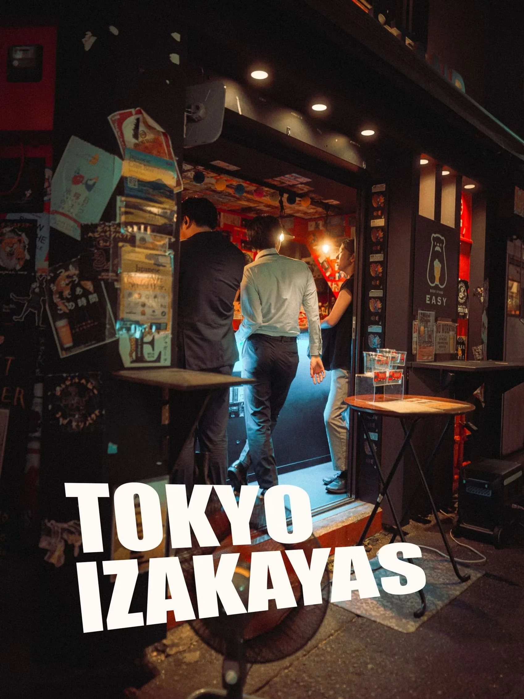 Night scene with a Japanese food cart decorated with Izakayas signs, people standing at the cart, and a large illuminated "TOKYO" sign in the foreground.