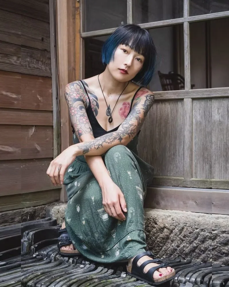 Young woman with black and blue hair, tattoos on her arms, sitting outside on a wooden porch near a rustic wooden building, wearing a black tank top, green patterned pants, and black sandals.