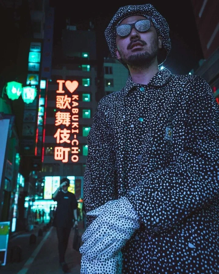 A man in polka-dot attire with neon signs and lights reflecting in the background, captured with an ultra-wide lens.