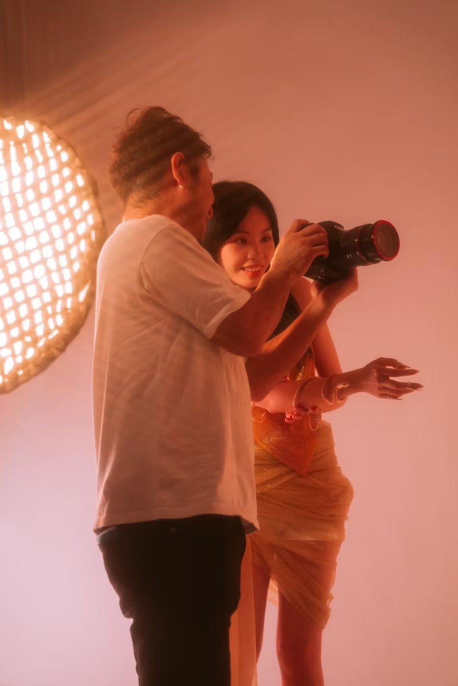 A photographer captures a model holding a bouquet of flowers while she poses gracefully in front of a large light. The setting is warm, with soft, flattering lighting that accentuates the model's pose and the natural beauty of the scene.