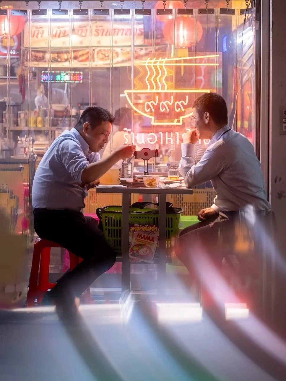 Two men in white shirts are sitting at a small table inside a restaurant, sharing a meal. The restaurant has a neon sign of a steaming bowl of food in the window behind them, with some Thai script visible. They are engaged in conversation.
