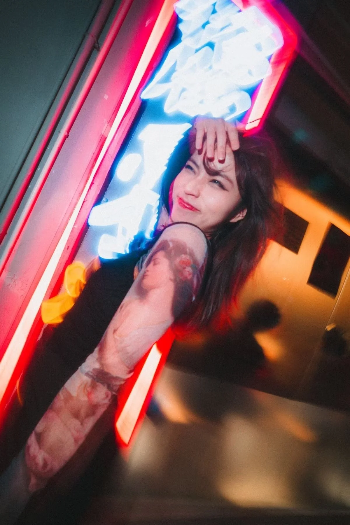 A woman with tattoos on her right arm, dark hair, and red lipstick, smiling and posing in front of a neon sign with blue, red, and white lights. Shot in Hong Kong by Deniz Demir.