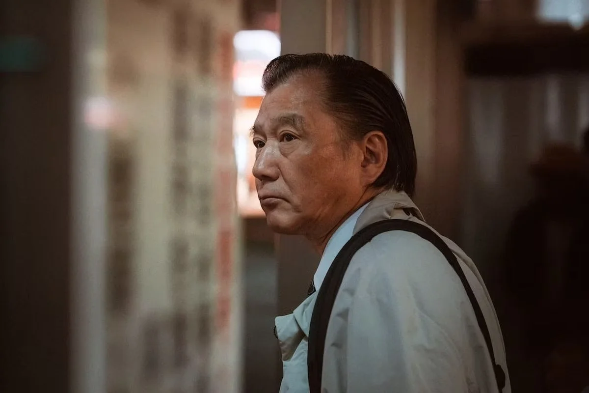 A middle-aged Japanese salaryman with slicked-back hair looking pensively to the side in a dimly lit indoor space.