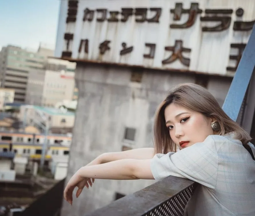 A woman with shoulder-length brown hair leaning on a balcony railing with a cityscape in the background.
