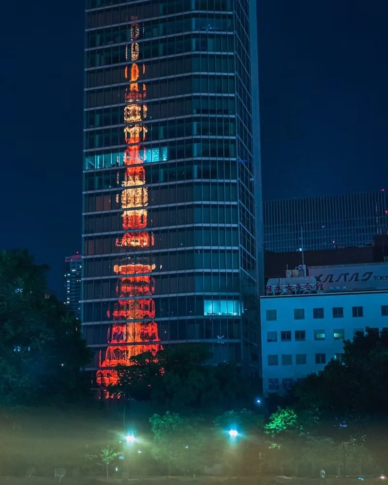 Nighttime cityscape with a tall glass building reflecting the illuminated Tokyo Tower, which is lit in red and orange lights, in the building's glass facade.